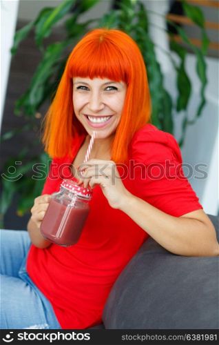 Happy woman drinking a blueberry smoothie in a glass jar with a straw at home