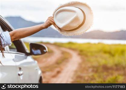 Happy woman hand holding hat outside open window car with meadow and mountain lake background. People lifestyle relaxing as traveler on road trip in holiday vacation. Transportation and travel concept