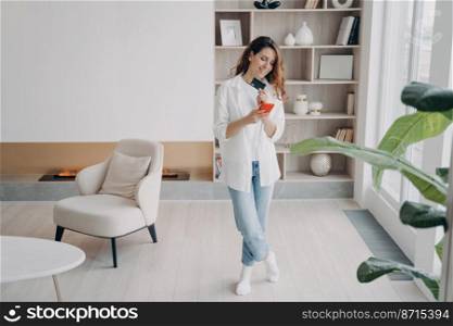 Happy woman holding bank credit card, smartphone, purchasing using online store app. Female homeowner tenant making cashless payment for house utilities, standing in modern living room at home.. Female holds bank credit card, phone makes cashless payment for house utilities in modern apartment