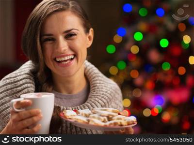 Happy woman holding cup of hot chocolate with marshmallows and plate of Christmas cookies