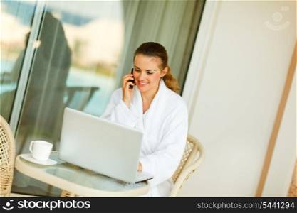Happy woman in bathrobe sitting at table on terrace speaking mobile and looking in laptop