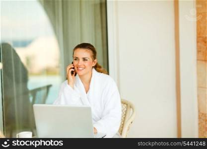 Happy woman in bathrobe sitting at table on terrace with laptop and speaking mobile
