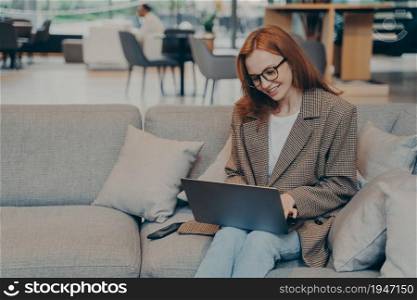 Happy woman in glasses sitting on cozy couch in lounge, using laptop to chat with family, satisfied with workplace environment, successful red haired business woman working remotely in coworking space. Woman in glasses sitting on cozy couch in office lounge while chatting on computer laptop