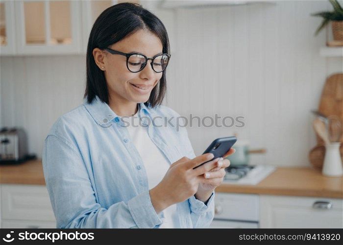 Happy woman in glasses uses mobile apps, shops online in kitchen. Smiling, she checks news, chats on social networks.