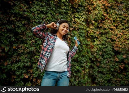 Happy woman in headphones listening to music in summer park. Female music fan walking outdoors, girl in earphones, green bushes on background. Woman in headphones listening to music in park