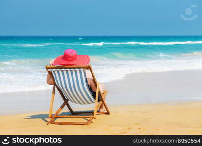 Happy woman in red sunhat on the beach sitting on deckchair and looking into the sea.Vacation and travel concept.