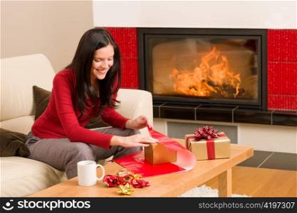 Happy woman in red wrapping Christmas present by home fireplace
