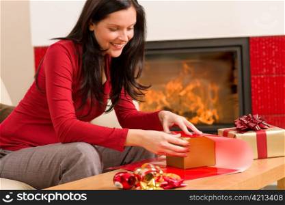Happy woman in red wrapping Christmas present by home fireplace