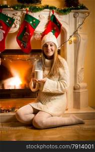 Happy woman in warm sweater and hat sitting at fireplace with tea cup