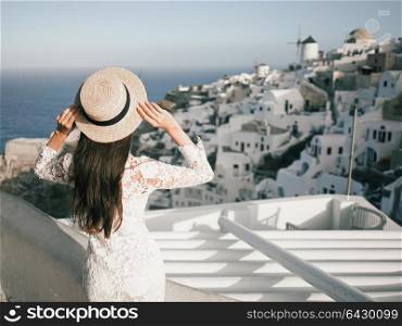 Happy woman in white dress and straw hat enjoying her holidays on Santorini island. View on Aegean sea from Oia. Europe summer travel destination. Greek Islands