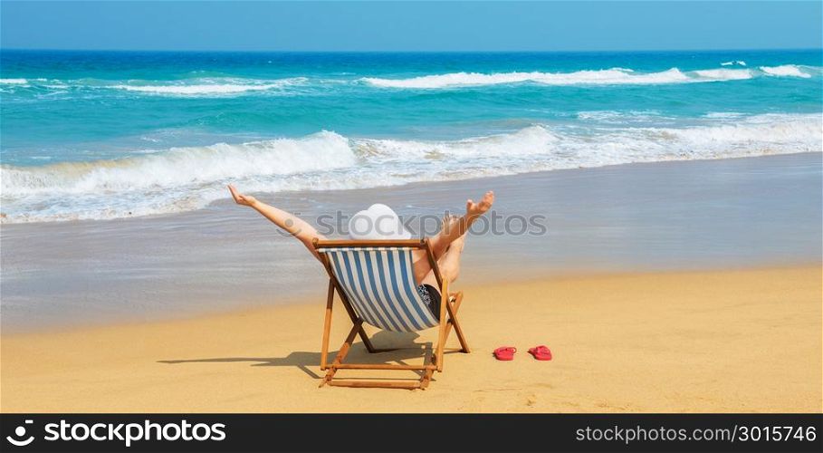 Happy woman in white sunhat on the morning beach sitting on deckchair with hands up.Vacation and travel concept.