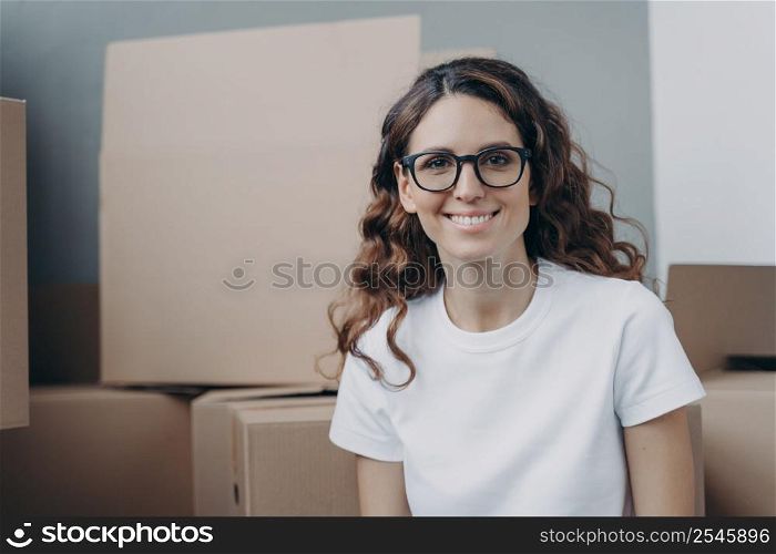Happy woman is an apartment buyer. Satisfied spanish girl is sitting among boxes in living room of new home. Young manager or freelancer is moving. Cheerful face of lady in glasses.. Satisfied spanish girl is sitting among boxes in living room of new home. Young woman is moving.
