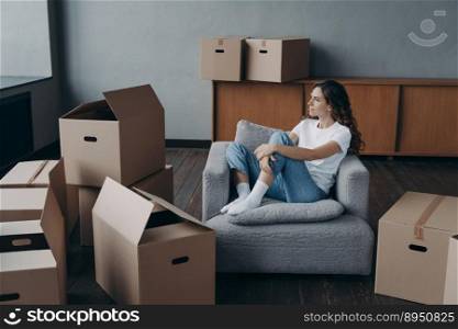 Happy woman is an apartment buyer. Satisfied spanish girl is sitting in armchair. Mover is dreaming among boxes in living room of new home. New house owner having rest.. Happy woman is an apartment buyer. Mover is dreaming among boxes in living room of new home.