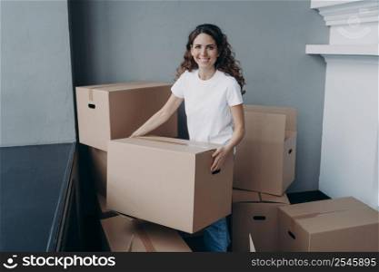 Happy woman is carrying box and smiling. Lady is purchasing real estate and unpacking cardboard boxes in new apartment. Young woman is going to rent house. Changes and new life concept.. Happy woman is carrying box and smiling. Lady purchasing real estate and unpacking cardboard boxes.