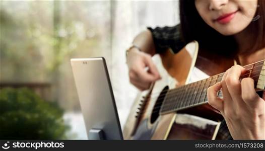 Happy Woman Learning an Acoustic Guitar in House via Tablet at Home. E-learning, Online Education Concept.Selective focus on a front hand