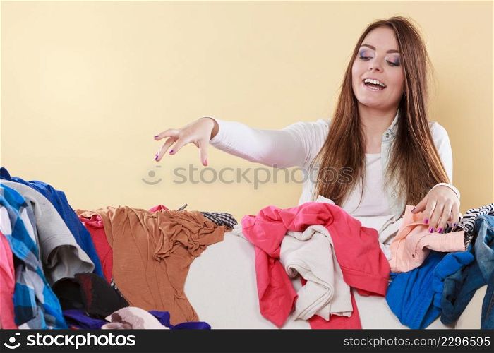 Happy woman picking clothes up off sofa couch in messy living room. Young girl surrounded by many stack of clothing. Disorder and mess at home.. Happy woman picking clothes up in messy room.