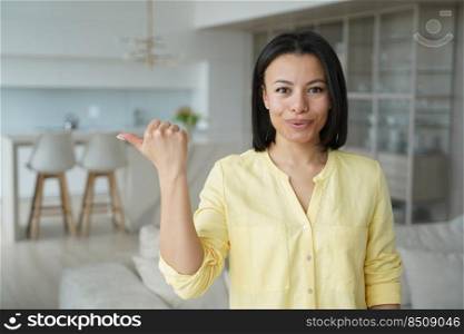 Happy woman points with thumb to copy space. Conceptual portrait of young emotional woman for advertising banner mockup. Handsome european girl in living room at home. Modern interior of apartment.. Happy woman points with thumb to copy space. Conceptual portrait of girl for advertising banner.