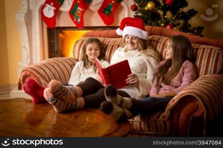 Happy woman reading book to daughters on sofa at fireplace at Christmas