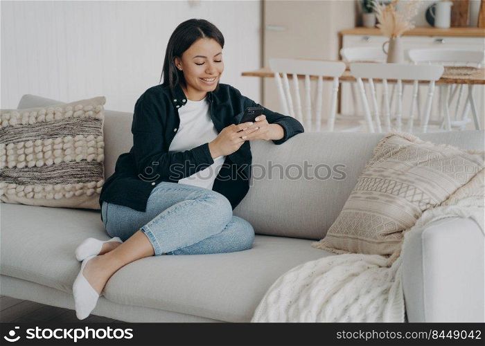 Happy woman relaxing with mobile phone on comfortable couch in living room. Girl texting and smiling, reading messages. Arab lady enjoying time at home in her apartment. Technology and internet using.. Happy woman relaxing with mobile phone and reading messages on comfortable couch in living room.