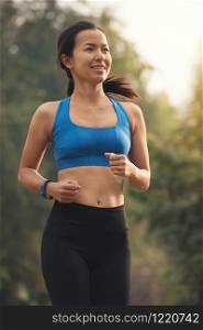 Happy woman running in outdoor park. Asian girl runner jogging smiling aspirational outside on beautiful summer day. female fitness sport model blue cloth black trouser with smart watch training outdoors.