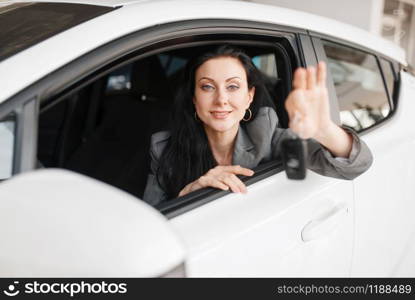 Happy woman shows the key to the new car in showroom. Female customer buying vehicle in dealership, automobile sale