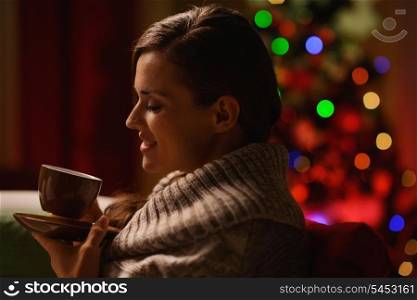 Happy woman sitting chair and drinking hot beverage in front of Christmas tree