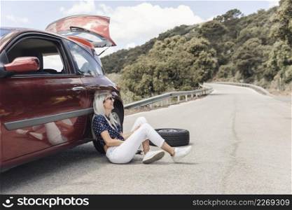 happy woman sitting near broken down car winding road
