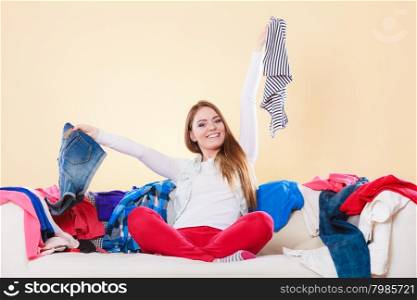 Happy woman sitting on sofa in messy room at home.. Happy woman sitting on sofa couch in messy living room holding clothes. Young girl surrounded by many stack of clothing. Disorder and mess at home.