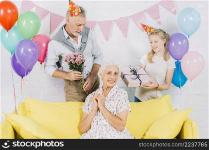 happy woman sitting sofa front husband granddaughter holding birthday gifts