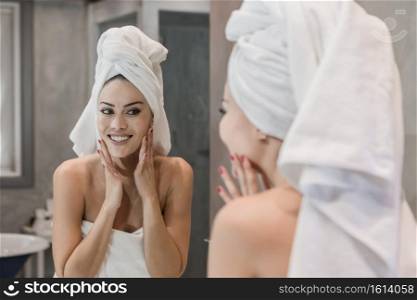 Happy woman smiling and touching clean skin of face while looking at mirror and applying cream after shower in bathroom. Cheerful female touching face after shower