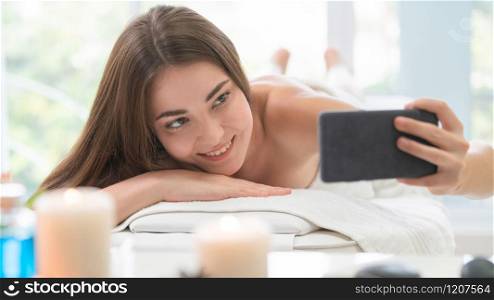 Happy woman takes selfie photo while lying on spa bed for aromatherapy massage in luxury spa with blurred foreground of spa treatment set. Wellness and healing concept.. Woman lying on spa bed for massage in luxury spa.