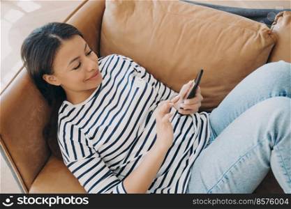 Happy woman using modern mobile apps on smartphone lying on sofa at home. Female housewife holding phone, shopping in online store, checking social networks news on couch. E-commerce concept.. Happy woman using modern mobile apps on smartphone, shopping in online store lying on sofa at home