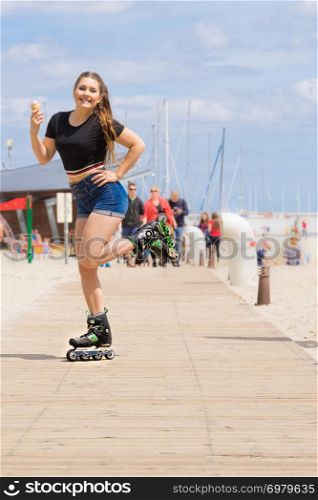 Happy woman wearing roller skates enjoying her free time while eating ice cream during summer sunny weather on beach. Roller skate woman eating ice cream