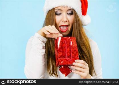 Happy woman wearing santa claus hat looks into christmas red gift bag, on blue. Christmas Woman looks into red gift bag