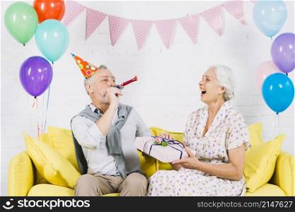 happy woman with birthday gift looking her husband blowing party horn