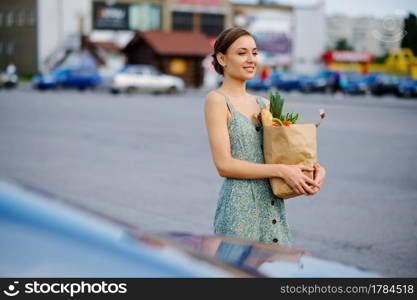 Happy woman with cardboard bag on supermarket car parking. Happy customers carrying purchases from the shopping center, vehicles on background. Happy woman with bag on supermarket car parking