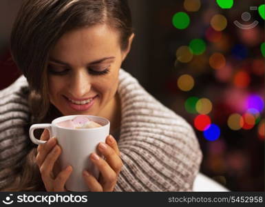 Happy woman with cup of hot chocolate with marshmallow in front of christmas lights