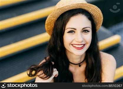 Happy woman with dark luxurious hair, charming bright eyes, well-shaped red lips and pure skin touching with hand her hair, wearing summer hat, smiling pleasantly into camera while being outdoor