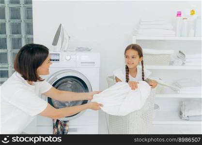 Happy woman with dark short hair pulls off laundry from basket, happy child poses in it, spend time in bathroom near washing machine and iron on top, shelf with folded white towels. Laundry time