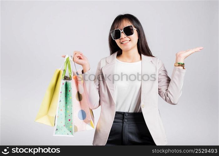 Happy woman with glasses shopper smile hold online shopping bags multicolor and show hand something empty for present product, excited Asian female purchase studio shot isolated on white background