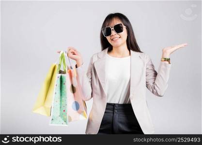 Happy woman with glasses shopper smile hold online shopping bags multicolor and show hand something empty for present product, excited Asian female purchase studio shot isolated on white background