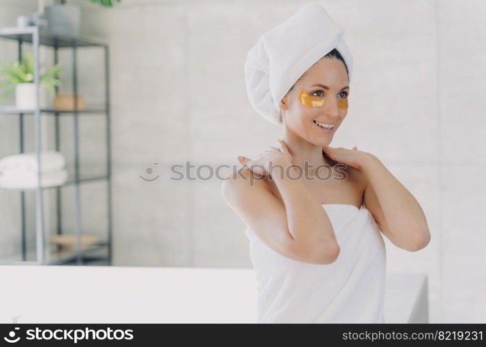 Happy woman with golden hydrogel under eye patches on face skin. Female in white bath towel on head and naked shoulders enjoying daily anti wrinkle skincare routine in bathroom after morning shower.. Woman with hydrogel eye patches on face skin enjoying skincare routine in bathroom after shower