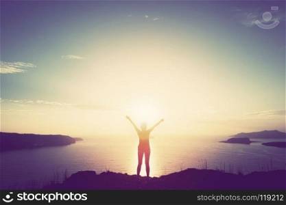 Happy woman with hands up standing on cliff over sea and islands at sunset. Vintage mood, concepts of winner, freedom, happiness etc.