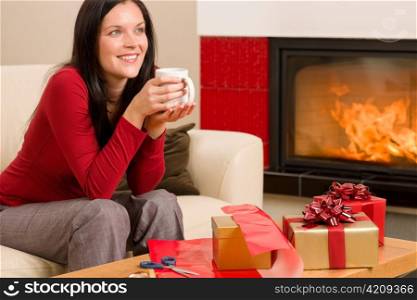 Happy woman wrapping Christmas present by fireplace enjoying hot drink