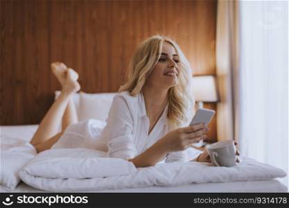 Happy womanl using a mobile phone while lying on the bed and drink morning coffee at home