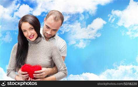 Happy young adult couple with red heart on romantic background with sky and clouds, embracing and laughing. Lots of copyspace
