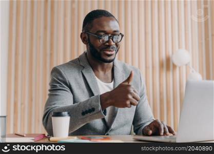 Happy young african american male employee in glasses talks online on laptop with client or business partner, showing thumb up and smiling, cheerful dark-skinned businessman making video call at work. Happy young african american businessman talks online on laptop with client or business partner