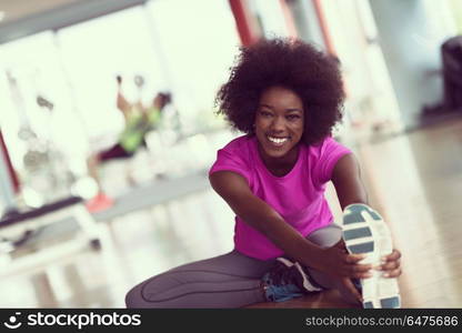 happy young african american woman in a gym stretching and warming up before workout young mab exercising with dumbbells in background. woman in a gym stretching and warming up man in background worki