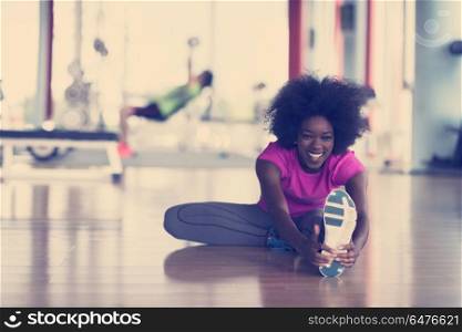 happy young african american woman in a gym stretching and warming up before workout young mab exercising with dumbbells in background. woman in a gym stretching and warming up man in background worki
