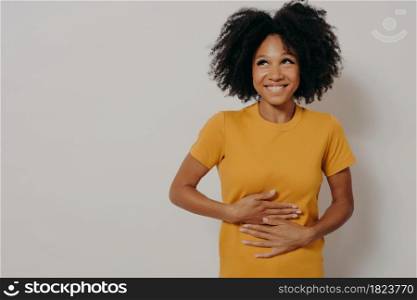 Happy young african american woman laughing out loud at some hilarious funny joke from friend, keeping both hands on stomach, looking aside cheerfully while standing isolated over white background. Happy african american woman laughing out loud at some hilarious joke, keeping hands on stomach